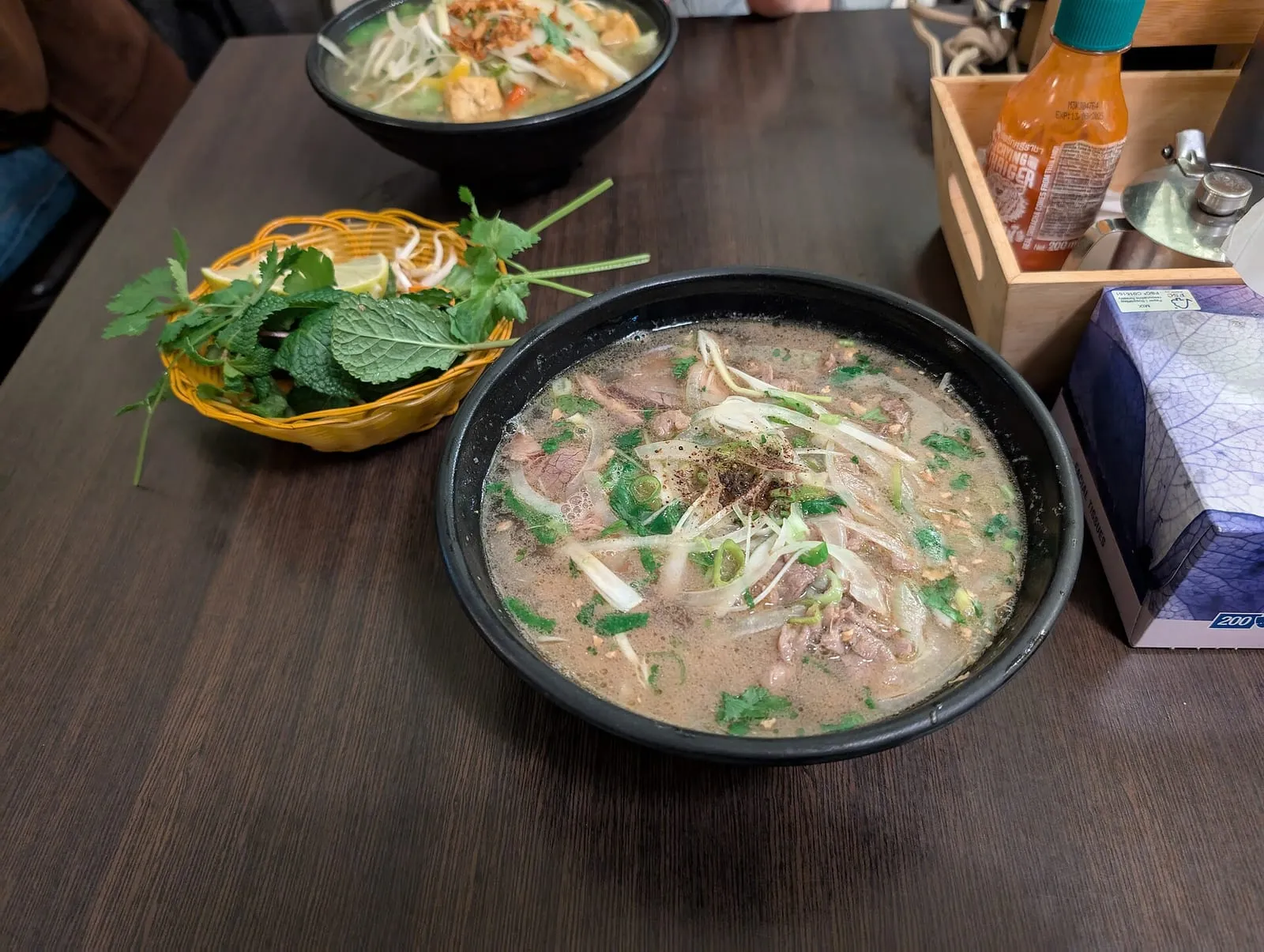 Schale Phở Bò im Restaurant, daneben ein Korb mit frischen Kräutern