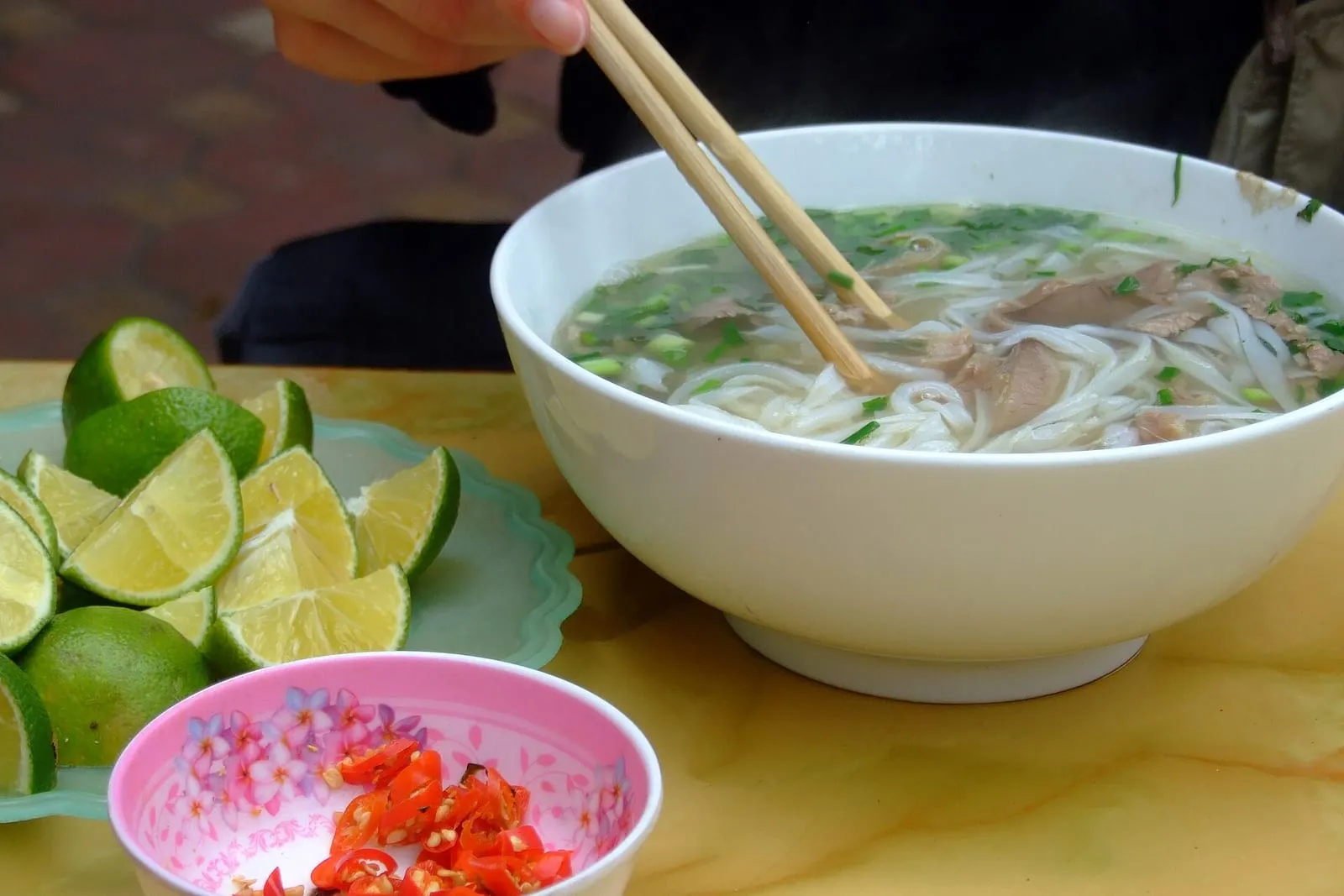 Phở Bò in einer großen weißen Schale, daneben Limetten und Chilis